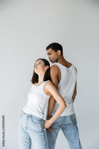 sensual asian woman with closed eyes leaning on african american man in white tank top and jeans isolated on grey.