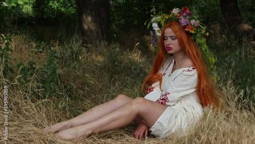 Fairytale character. Beautiful girl with long red hair with flower crown in embroidered ethnic suit sits in forest among trees. Paganism. Forest spirit, mavka, fairy. 4K Resolution