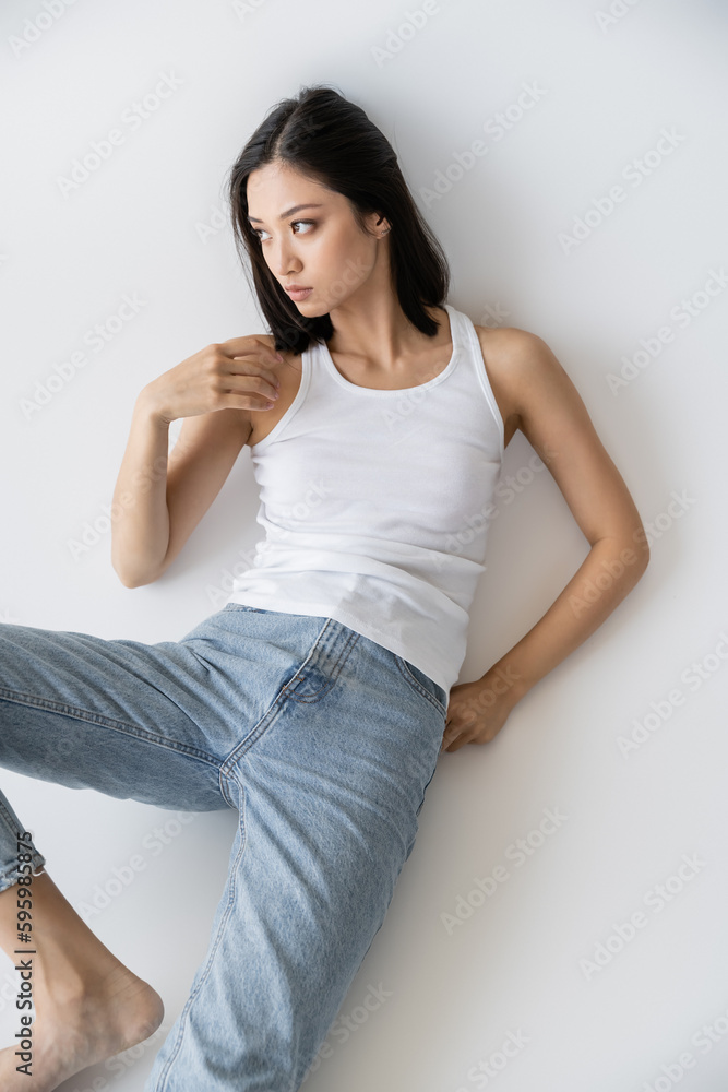 high angle view of asian woman in white tank top and jeans looking away on grey background. high angle view of asian woman in white tank top and jeans looking away on grey background.