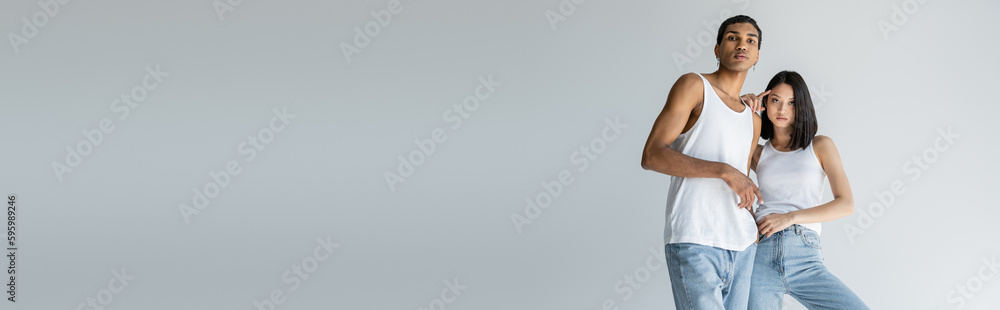 Naklejka premium young multiethnic couple in white tank tops and jeans looking at camera isolated on grey, banner.