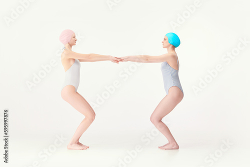 Two young girls, swimming athletes in retro style swimsuits standing in jump position isolated over white background. Training. Concept of retro style, sport, fashion, youth, vintage
