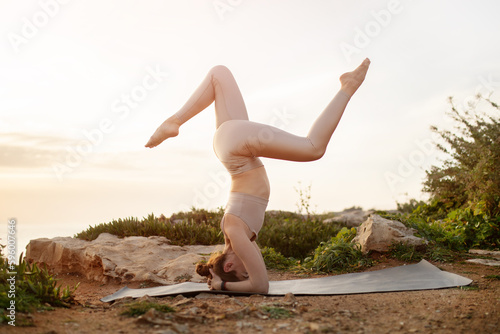 Smiling young caucasian lady in sportswear practicing yoga on head on ocean beach at sunrise, full length, sun flare