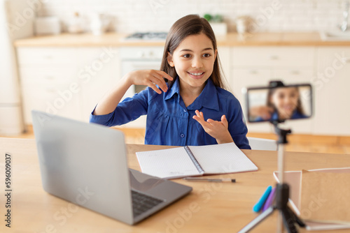 Schoolgirl Video Calling Via Smartphone Sitting At Laptop Learning Indoors © Prostock-studio