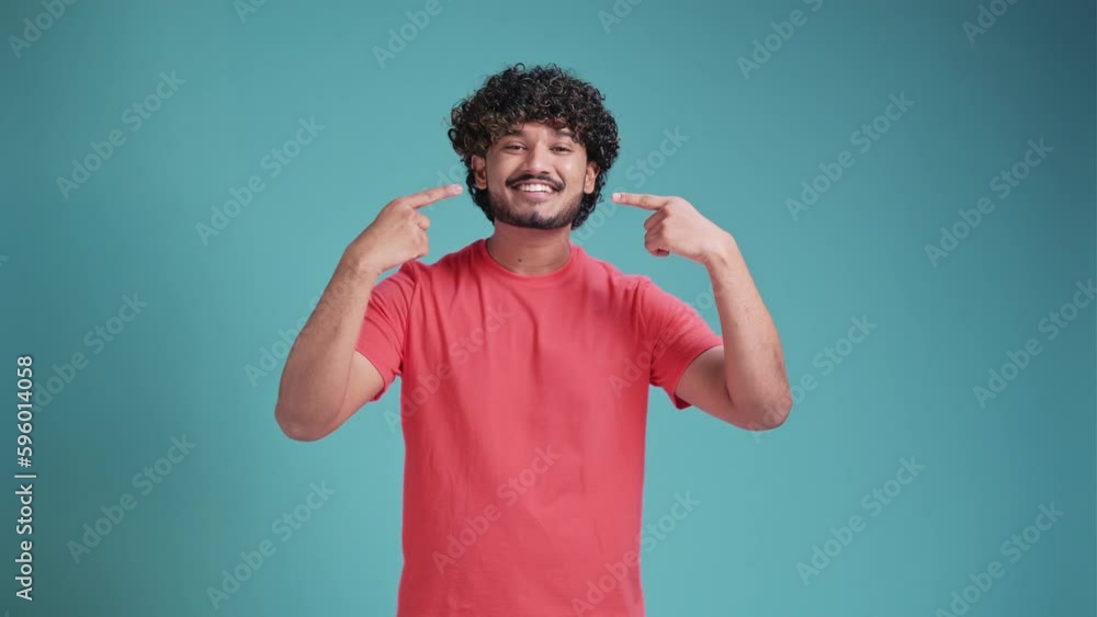 young hispanic man smiling and pointing at his teeth and beautiful smile on blue background young hispanic man smiling and pointing at his teeth and beautiful smile on blue background