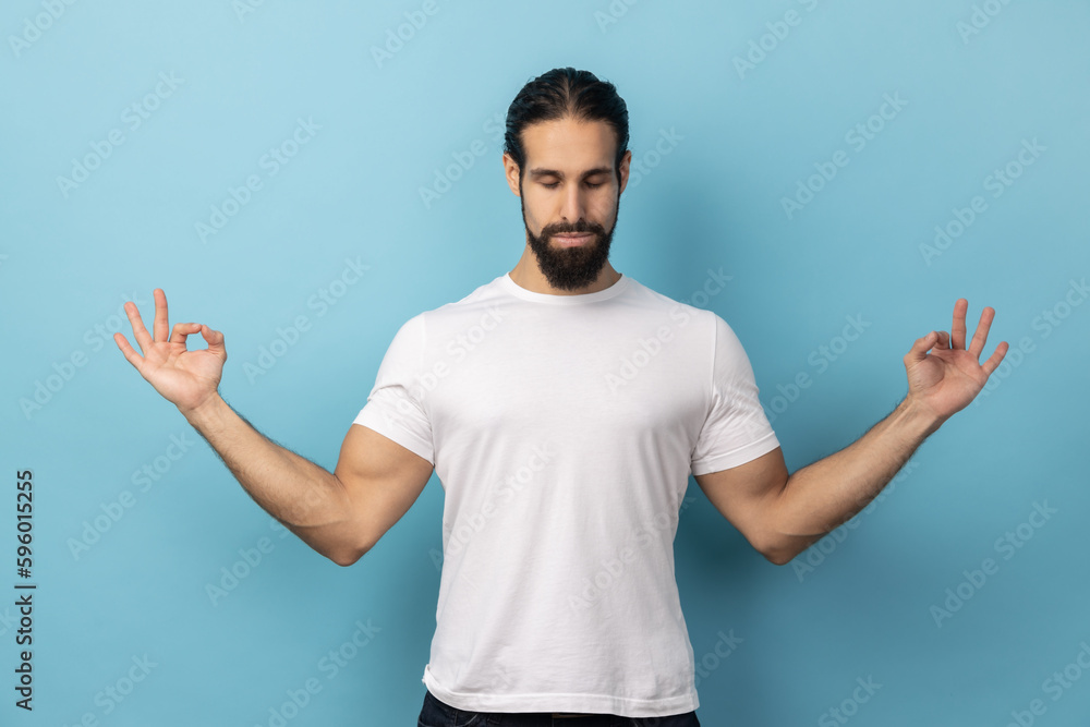 Portrait of calm relaxed man with beard wearing white T-shirt doing yoga, breathing, keeping eyes closed, calming down after work. Indoor studio shot isolated on blue background. Portrait of calm relaxed man with beard wearing white T-shirt doing yoga, breathing, keeping eyes closed, calming down after work. Indoor studio shot isolated on blue background.
