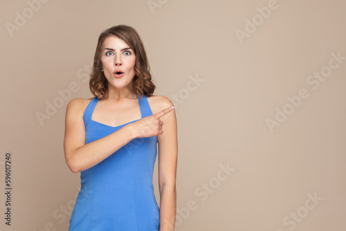 Portrait of surprised woman with wavy hair showing something aside at blank wall, being astonished, advertisement area, wearing blue dress. Indoor studio shot isolated on light brown background.