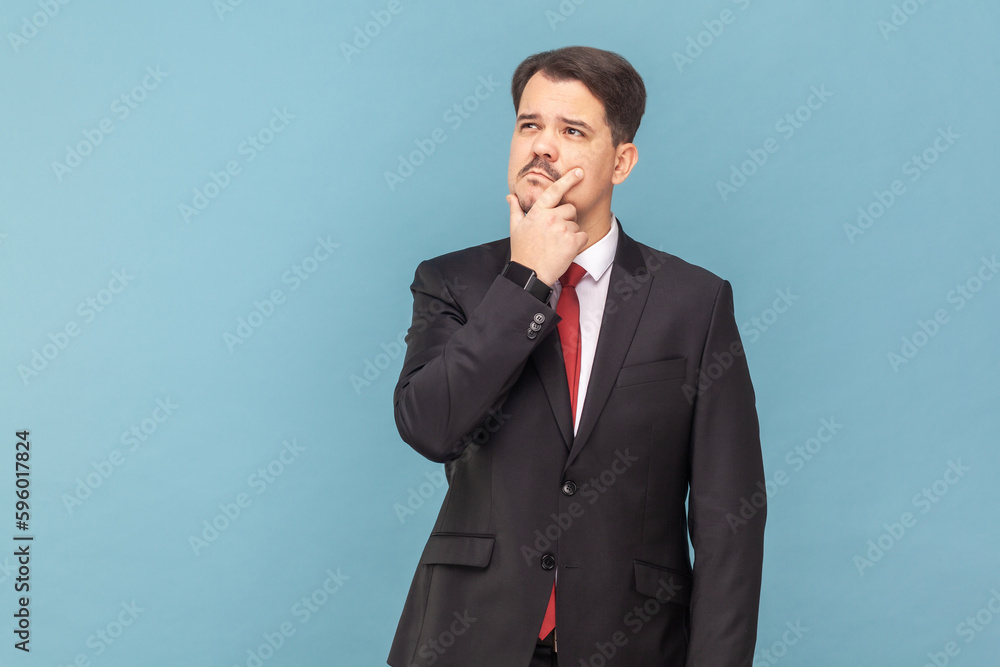 Portrait of pensive thoughtful man with mustache standing with puzzled expression, looking away, holding chin, wearing black suit with red tie. Indoor studio shot isolated on light blue background. Portrait of pensive thoughtful man with mustache standing with puzzled expression, looking away, holding chin, wearing black suit with red tie. Indoor studio shot isolated on light blue background.