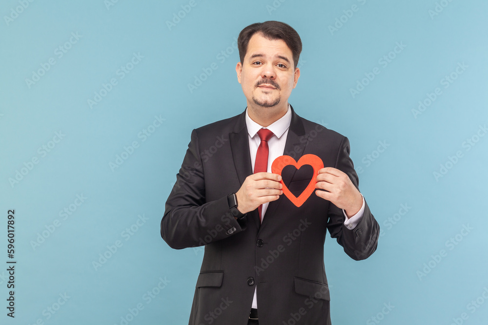 Portrait of attractive man with mustache standing holding red gesture, looking at camera with positive expression, wearing black suit with red tie. Indoor studio shot isolated on light blue background Portrait of attractive man with mustache standing holding red gesture, looking at camera with positive expression, wearing black suit with red tie. Indoor studio shot isolated on light blue background