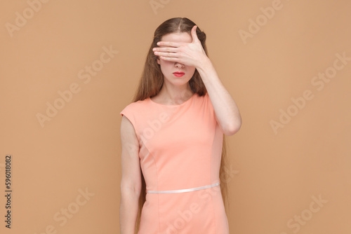 Portrait of serious sad woman with long hair standing and covering her eyes with her palms, doesn't want to see something shame, wearing elegant dress. Indoor studio shot isolated on brown background.