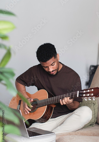 Cheerful ethnic man playing guitar with laptop on sofa