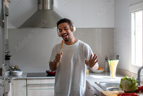 Cheerful ethnic man in headphones with spatula singing in kitchen photo