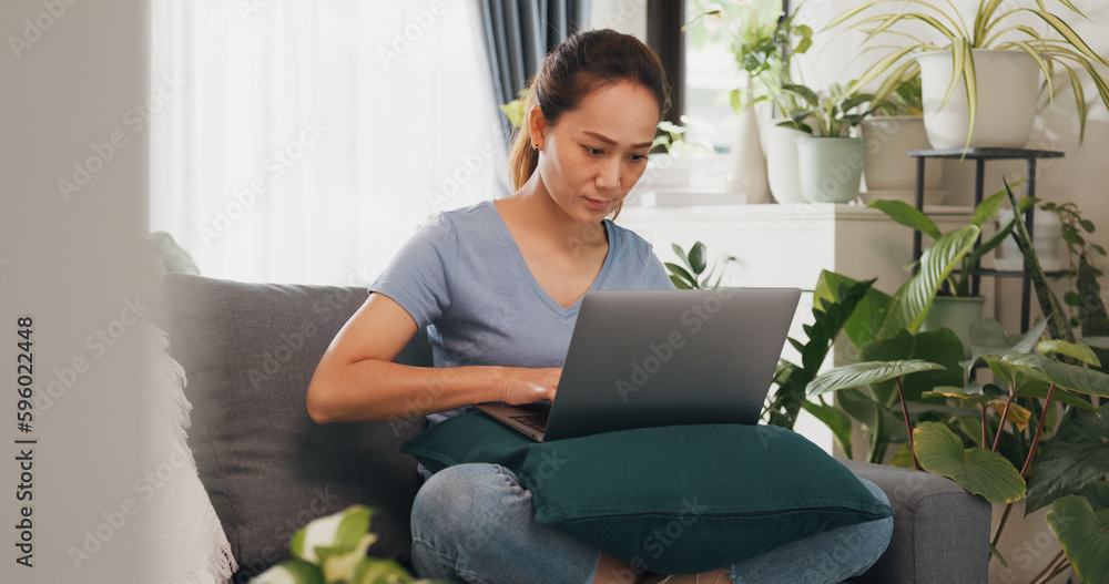 Young Asian woman wear casual calm sitting on wing chair use laptop computer thinking the idea for working full of plants in living room greenhouse. Coronavirus and stay home, Work from home concept. Young Asian woman wear casual calm sitting on wing chair use laptop computer thinking the idea for working full of plants in living room greenhouse. Coronavirus and stay home, Work from home concept.