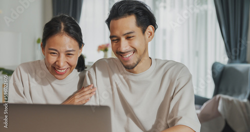 Happy young Asian couple sit on couch with laptop success credit card payment online shopping at home on holiday. Smile young husband and wife excited browsing app on gadgets. Lifestyle concept.