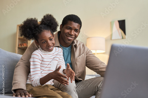 African American father sitting on sofa with his little daughter and using laptop for online communication