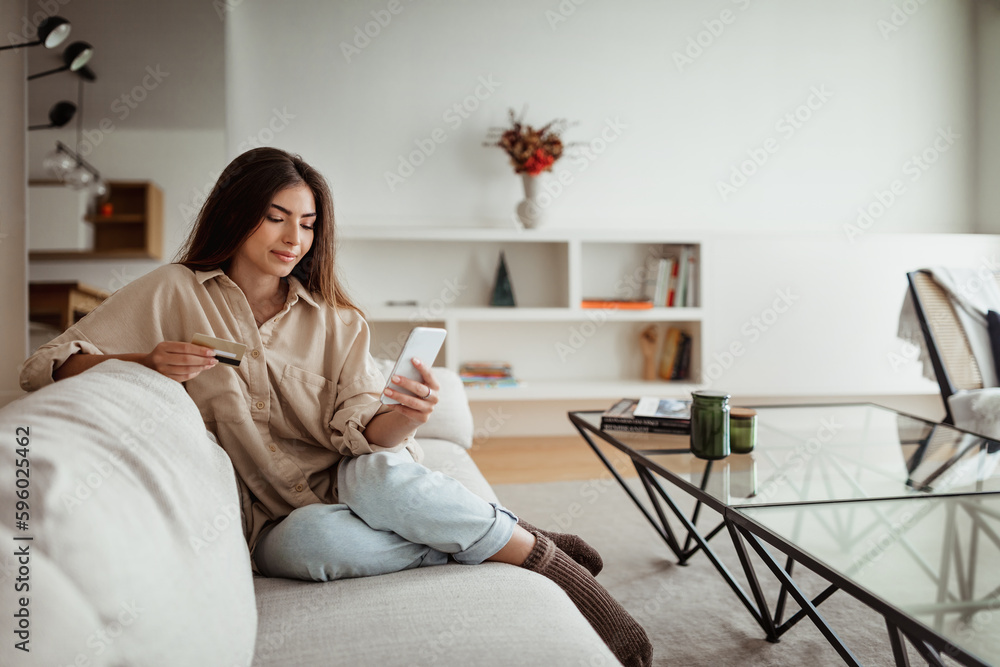 Positive millennial european lady shopaholic with credit card looking at phone, order purchases
