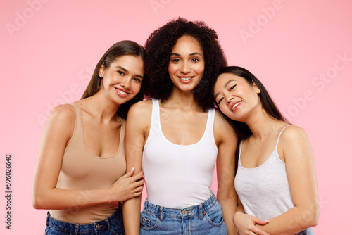 Happy multiethnic young women posing and smiling to camera, standing over pink background, studio shot