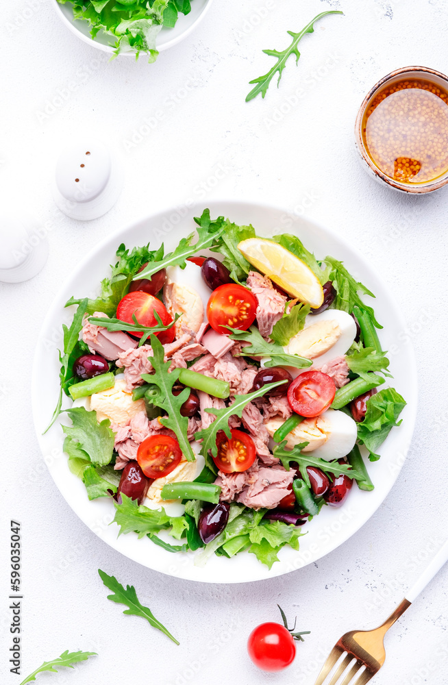 Nicoise salad with tuna, red tomatoes, boiled eggs, green beans and olives on plate, white table background, top view