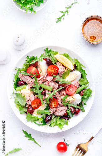 Nicoise salad with tuna, red tomatoes, boiled eggs, green beans and olives on plate, white table background, top view