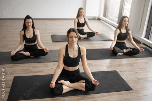 Group of attractive sporty women in black activewear sitting in lotus pose on sticky mats in spacious studio room. Padmasana in yoga calming mind and preparing practitioners for deep meditation.