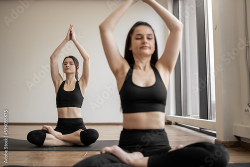 Attractive adult females with sports outfit and closed eyes holding folded palms overhead while meditating in crossed-legged position in fitness studio. Slim yogis practicing Padmasana together.