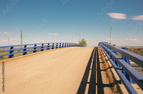 Overpass bridge against blue sky. Asphalt paved road, metal guardrails at midday. Travel in warm summer sunny day. Straight pathway goes over a horizon. No people. Rural street scene. Infrastructure.