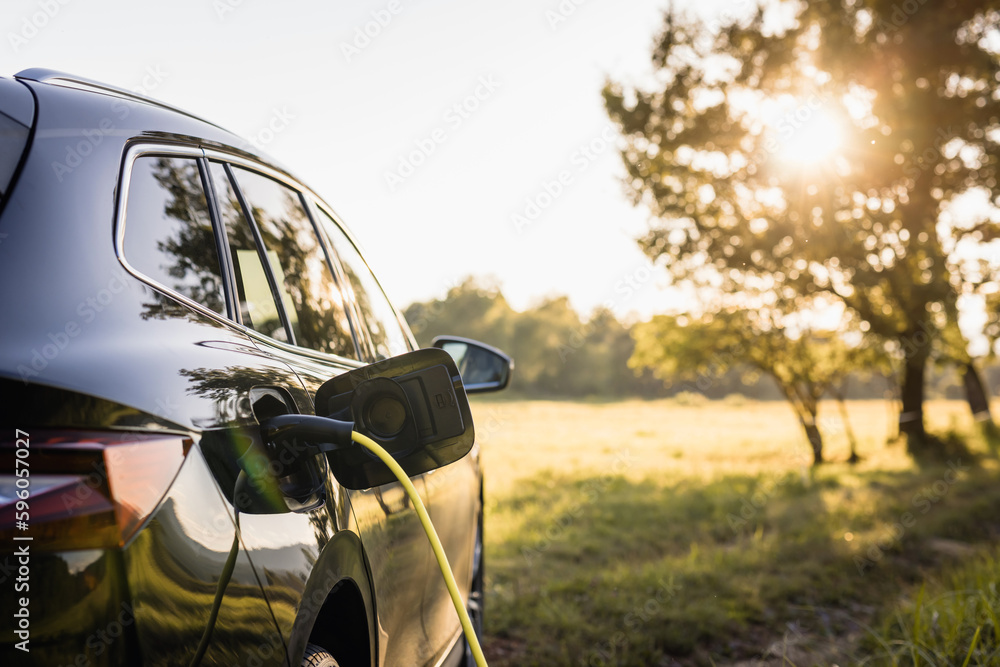 Black electric car parked in the nature area with a plugged cable and charging. Shining sun visible in background. Renewable energy concept. Black electric car parked in the nature area with a plugged cable and charging. Shining sun visible in background. Renewable energy concept.