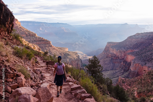 Rear view of woman with backpack hiking along Bright Angel trail with panoramic aerial overlook of South Rim of Grand Canyon National Park, Arizona, USA, America. Amazing vista after sunrise in summer