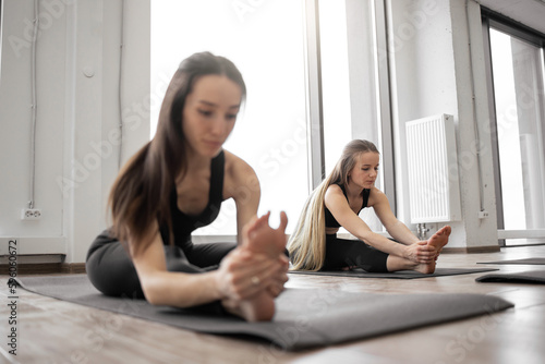 Young healthy females in sportswear doing Janu Sirsasana exercise on black mats in meditation room. Athletic caucasian yoginis eliminating mild depression while taking up Head to Knee Pose indoors. © sofiko14
