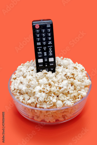 Popcorn in a glass bowl and a TV remote control on an orange vertical background. Leisure concept with watching movies or series and tasty snack in the form of crispy popcorn