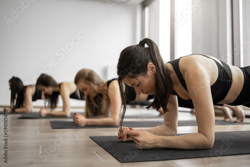 Close up view of attractive ladies in tank tops performing Phalakasana II on black mats indoors. Active yoga practitioners counteracting effects of prolonged sitting with help of Forearm Plank Pose.