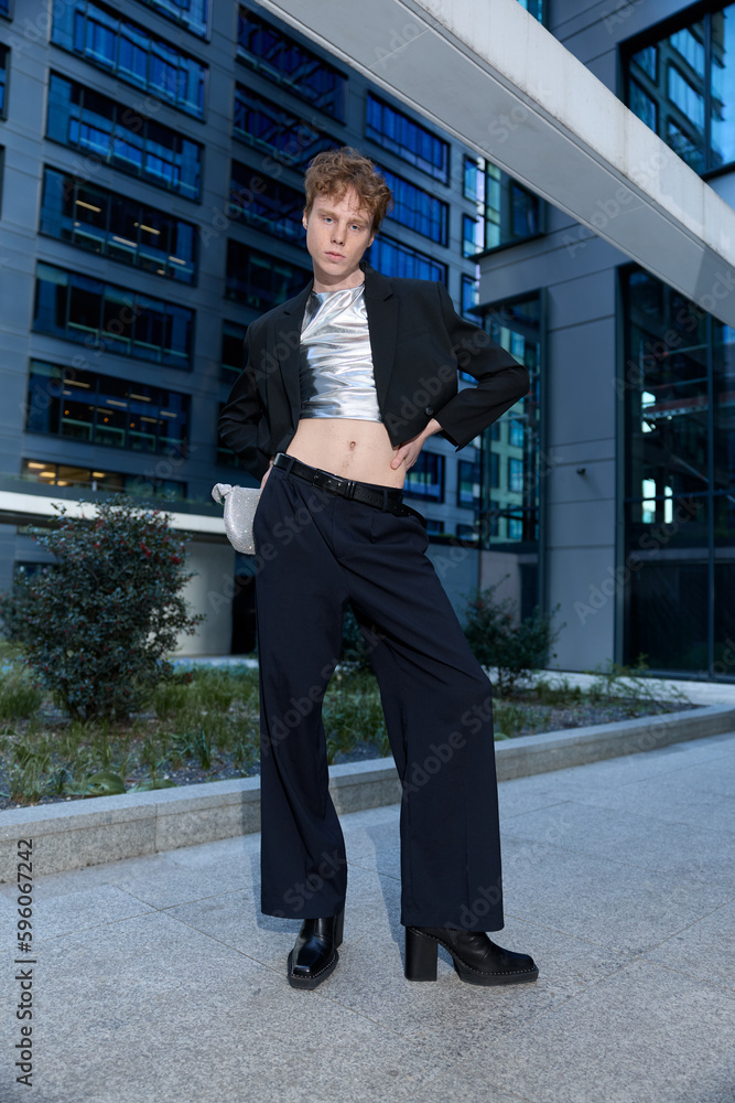 Full length vertical shot of ginger male fashion influencer model wearing elegant outfit black suit and heels standing in urban area against tall buildings and posing for photo with his hands on hips.