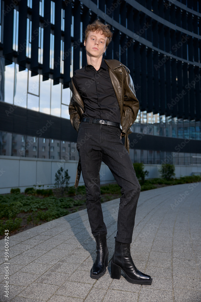 Vertical shot of fashionable male stylist influencer or designer wearing classy elegant outfit brown leather jacket, black trousers and black shirt with high heels cossack boots posing in urban area.