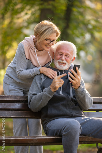 Happy senior couple relaxing together in a city park, sitting on a park bench and using a smart phone