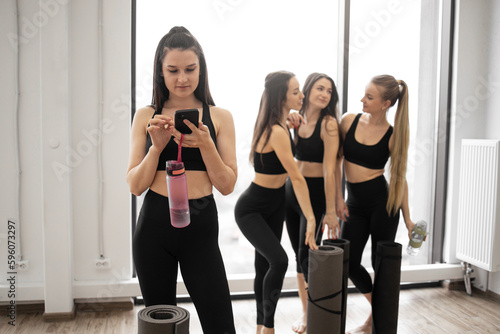 Beautiful brunette female in yoga outfit holding smartphone and sports bottle while standing with friends in fitness studio. Cheerful women chatting after full body workout in training room.
