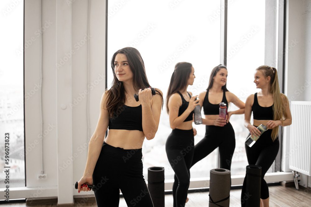 Fototapeta premium Beautiful slim brunette in activewear lifting dumbbells while fellow yoginis having rest after workout session. Young caucasian ladies with sports equipment talking in fitness studio after exercises.