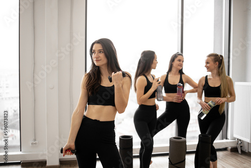 Beautiful slim brunette in activewear lifting dumbbells while fellow yoginis having rest after workout session. Young caucasian ladies with sports equipment talking in fitness studio after exercises.