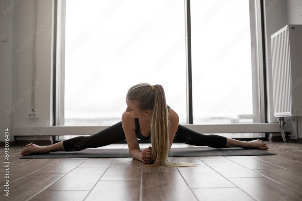 Beautiful blonde female in tight yoga clothing extending in Wide Angle Seated Forward Bend Pose on rubber mat in workout studio. Charming lady improving joint health with Upavistha Konasana posture.