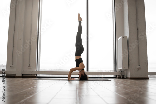 Concentrated strong female doing yoga while standing on her head in salamba sirsasana pose among brightly lit room. Athletic young woman in comfy sportswear strengthening her body and calming brain.