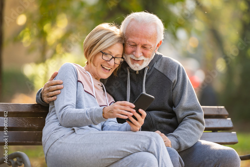 Happy senior couple relaxing together in a city park, sitting on a park bench and using a smart phone