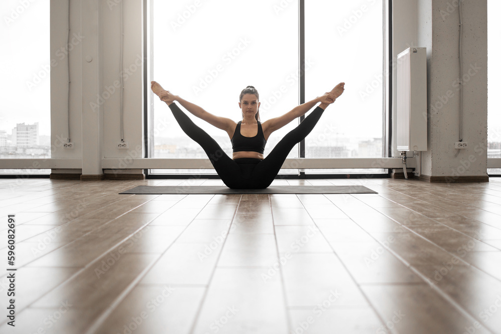 Front view of beautiful fitness model in black sportswear doing stretching while sitting on floor of white capacious studio. Caucasian dark haired lady balancing on yoga mat during everyday training.