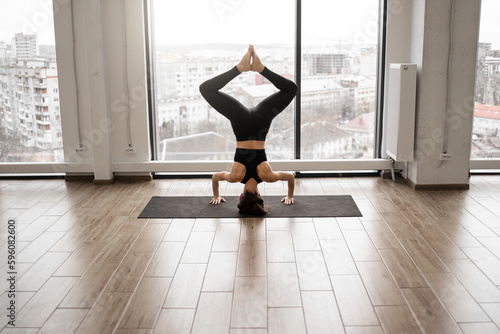Back view of strong caucasian woman with athletic body shape standing on hands and keepings legs clenched above head during yoga practice in studio. Panoramic windows on background.
