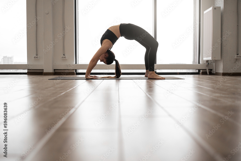 Obraz premium Attractive caucasian yogi dressed in black sport outfit standing in Urdhva Dhanurasana pose during workout. Active woman practicing Bridge exercise on yoga mat at room with french windows.