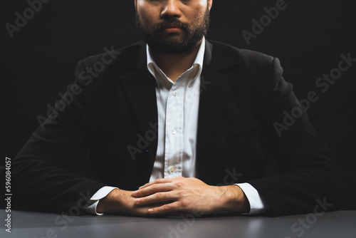 Businessman or lawyer wearing formal black suit sitting at table on isolated black background. Concept of a man with authority and seriousness gesture. equility photo