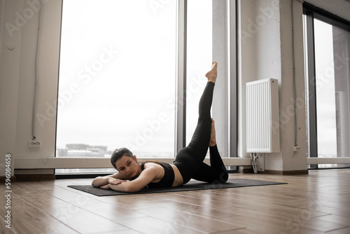 Side view of stunning young woman wearing black sportswear lying on mat and lifting legs up. Slim caucasian brunette practicing yoga pose at studio with panoramic windows.
