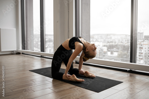 Athletic caucasian woman with bare feet standing in Camel Pose during training at fitness center. Flexible female yogi stretching in Ustrasana exercise with background of French windows.