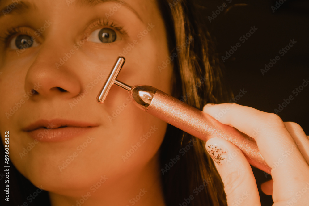 Obraz premium Young beautiful white woman, girl is using vibrating golden facial massager on dark black background with copy space. Preservation prolongation of natural beauty youth Female skin care with pleasure.
