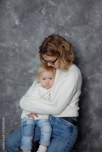 Portrait of family in white sweaters, jeans, sitting on grey background. Young beautiful woman embracing little girl.