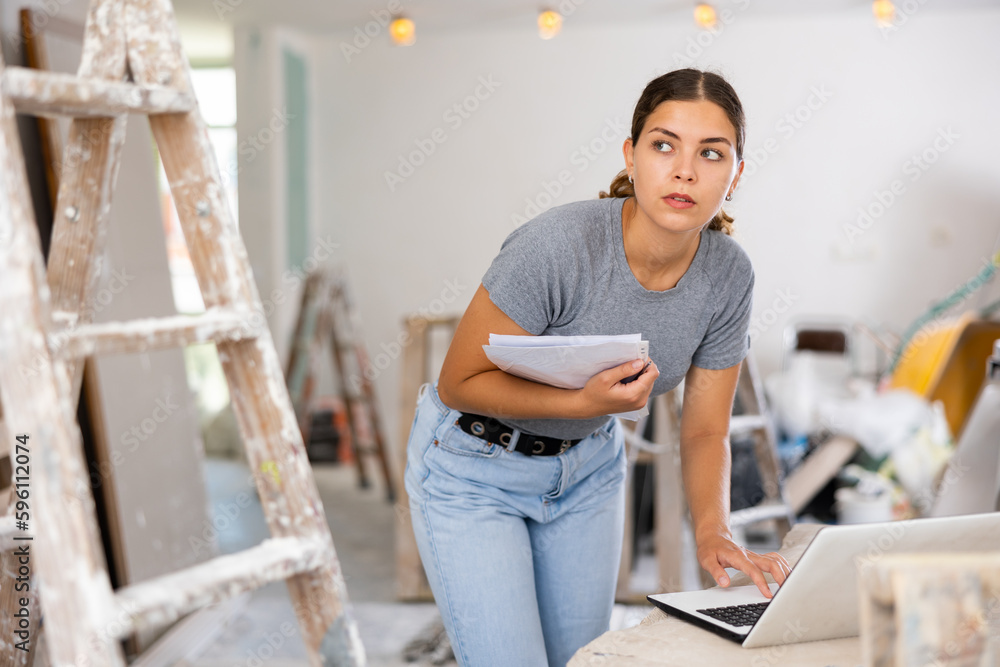 Young woman designer checking documents and using her laptop during repair works indoors.