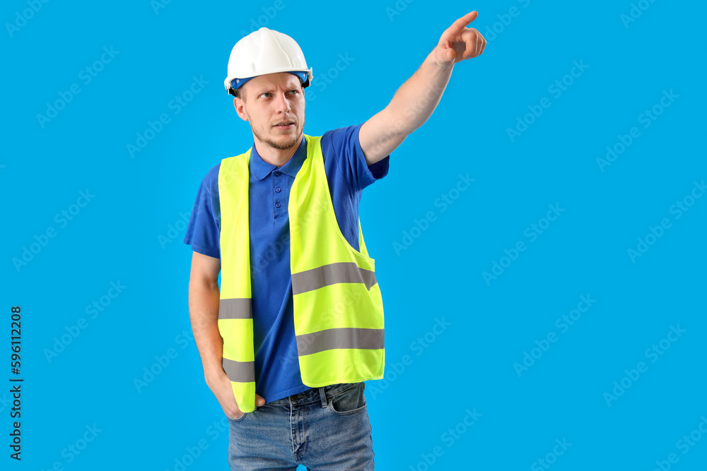 Male worker in vest and hardhat pointing at something on blue background Male worker in vest and hardhat pointing at something on blue background