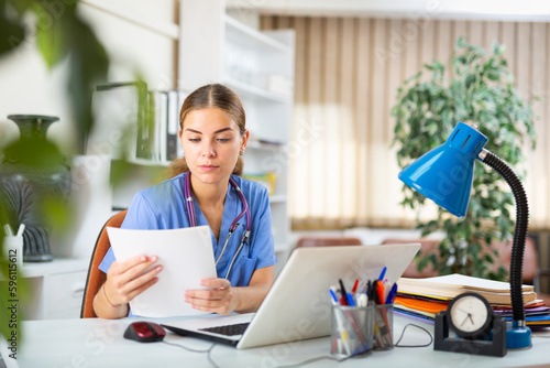 Female doctor in surgical scrubs sitting at desk in office and looking in documents in her hands.